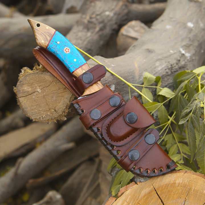 Knife with a blue blade and brown sheath on a wooden log with green leaves.